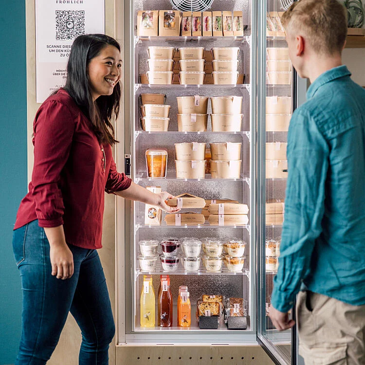 Eine junge Frau öffnet den gustav smart fridge und nimmt ein Menu heraus, während ihr ein junger Mann zusieht.