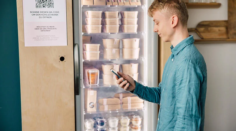 Ein junger Mann mit Smartphone in der Hand steht vor einem gustav smart fridge.